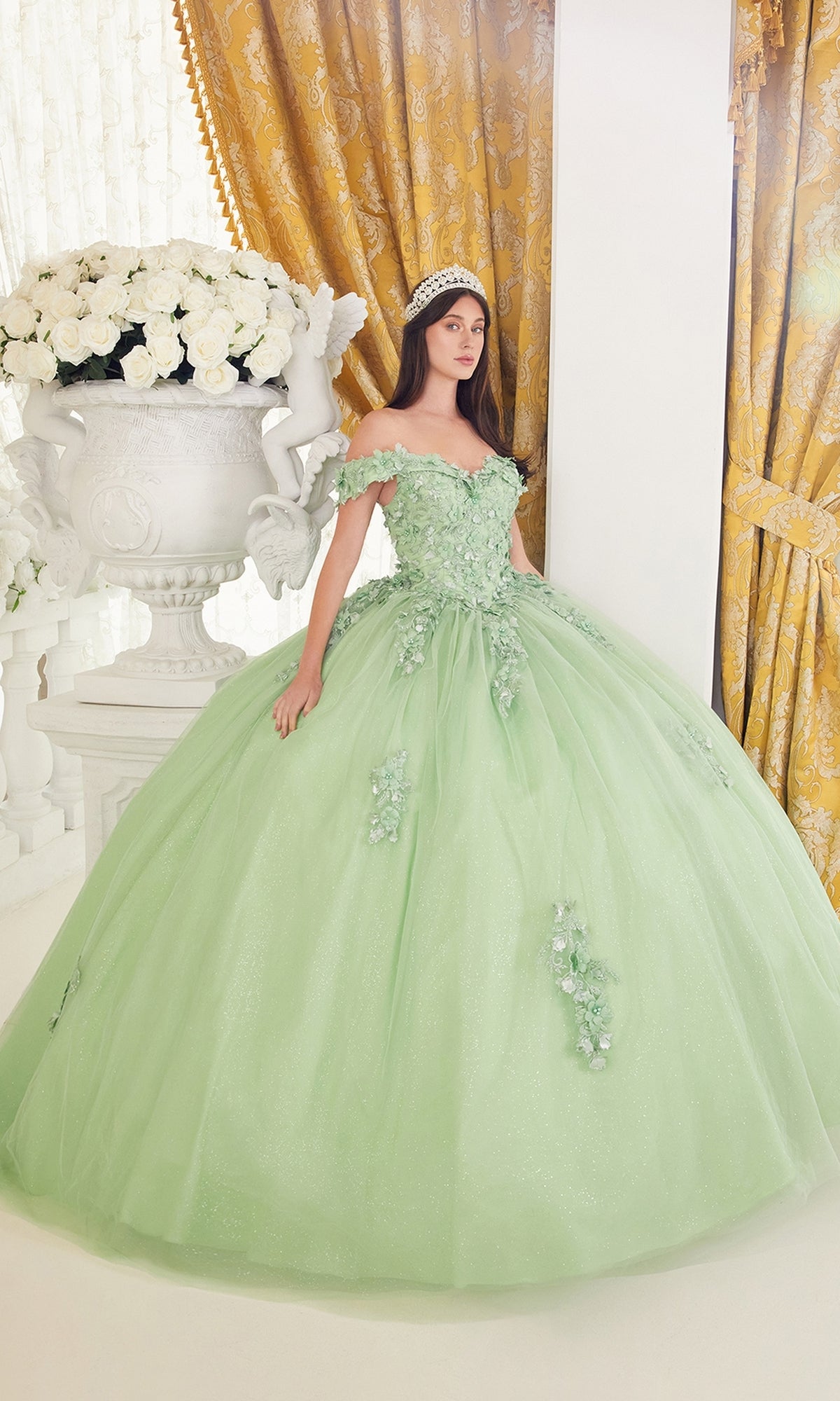 A young woman stands indoors beside a large white urn of roses, wearing the Ladivine 15710 Off-Shoulder Green Quinceañera Dress adorned with floral embellishments.